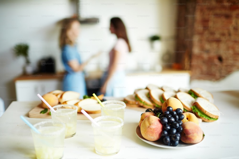 접시에 신선한 과일, 플라스틱 안경에 만든 수제 음료, 배경에 말하는 여자와 테이블에 샌드위치