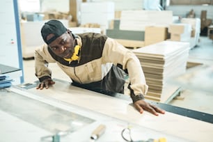 Serious African man in uniform controlling the process of sawing the wooden plank on machine at factory