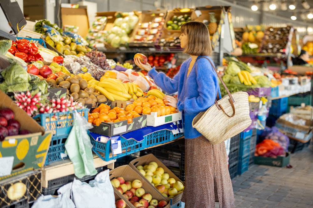 Giovane donna allegra che sceglie frutta fresca al mercato locale, facendo shopping con borsa a rete riutilizzabile. Sostenibilità e concetto di cibo biologico locale