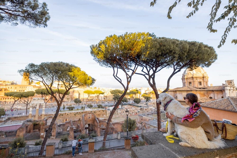 Mujer disfrutando de una hermosa vista desde arriba sobre el casco antiguo de Roma sentada junto con un perro. Mujer caucásica en vestido y chal en el pelo visitando el foro romano con el perro pastor italiano