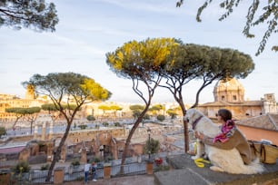 Mujer disfrutando de una hermosa vista desde arriba sobre el casco antiguo de Roma sentada junto con un perro. Mujer caucásica en vestido y chal en el pelo visitando el foro romano con el perro pastor italiano