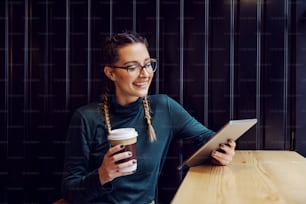Young smiling teenage geeky girl sitting in coffee shop , holding disposable cup with coffee and looking at tablet.
