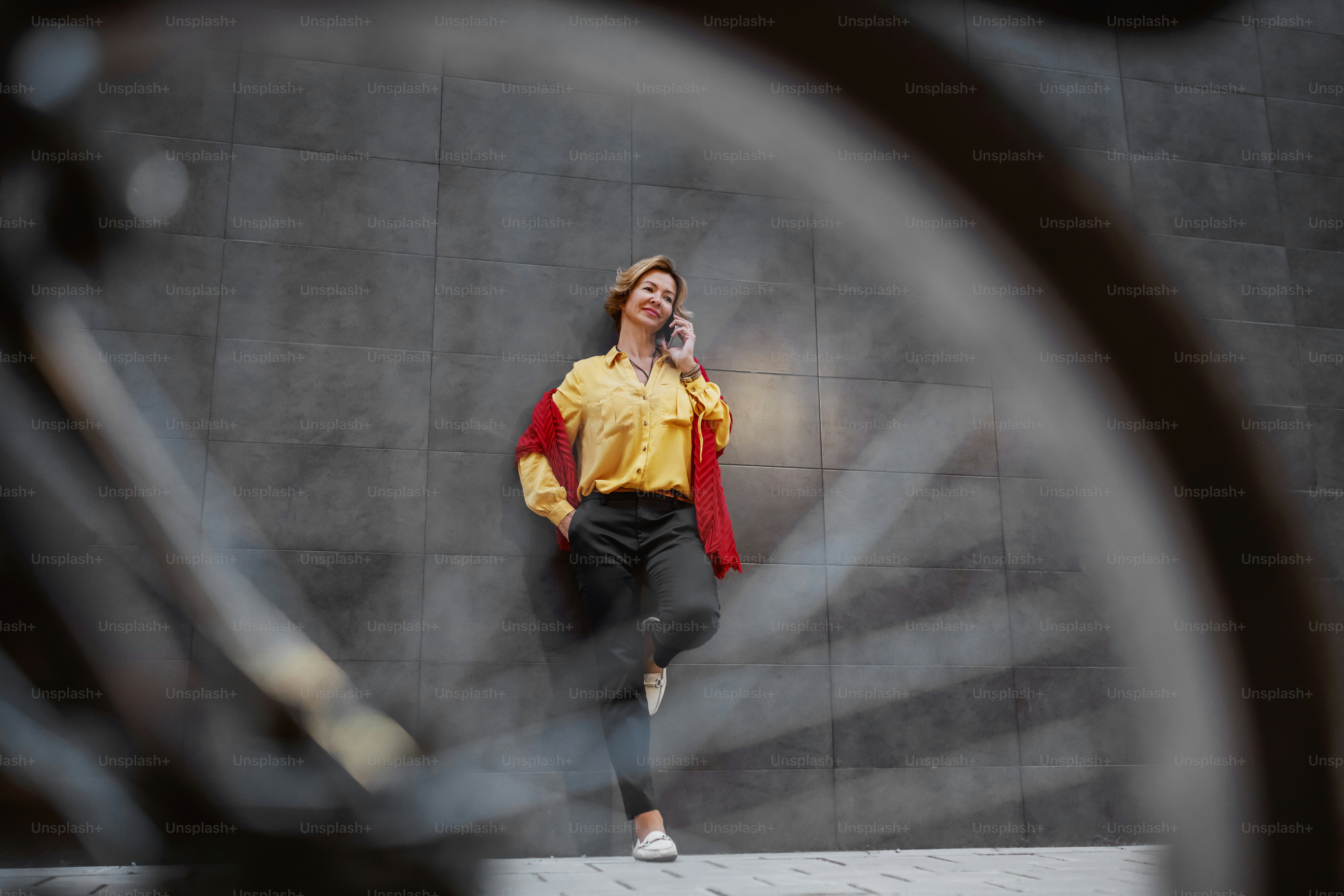 Full length of beautiful caucasian blonde fashionable senior woman with scarf talking on the phone while leaning on the wall outside. In foreground is bicycle.