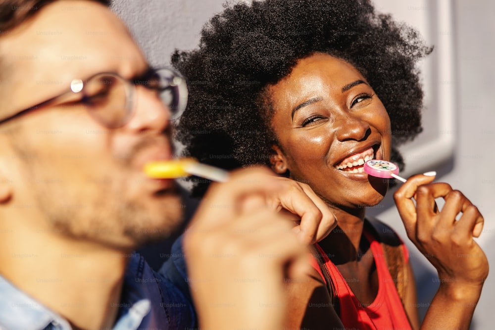 Young attractive multiracial couple leaning on the wall and licking lollypops.
