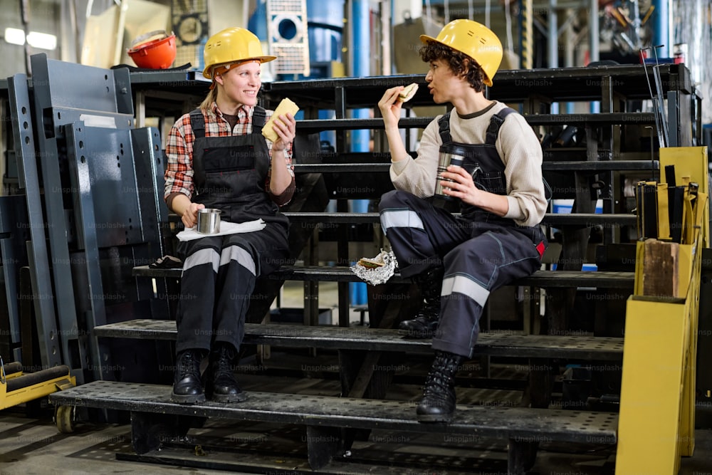 Two young female engineers chatting while sitting on staircase in distribution warehouse and having sandwiches with tea for lunch