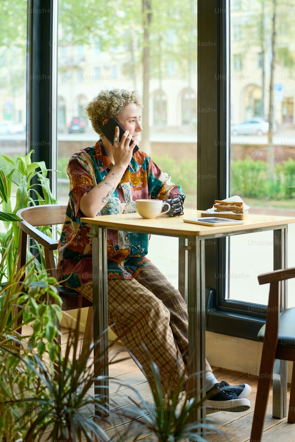Mujer joven con brazo parcial hablando por teléfono móvil junto a la mesa en la cafetería y mirando a través de una gran ventana mientras se relaja en el tiempo libre