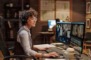 Giovane colorista in occhiali da vista che si siede sulla sedia al tavolo e lavora al computer con il software multimediale in ufficio