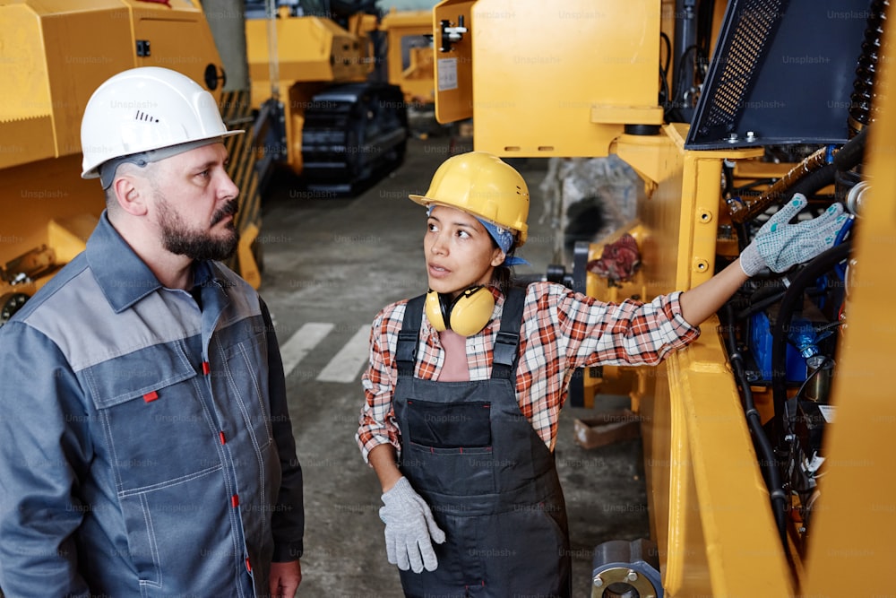 Mujer joven segura de sí misma en ropa de trabajo señalando el motor roto de una máquina industrial mientras consulta con su colega masculino
