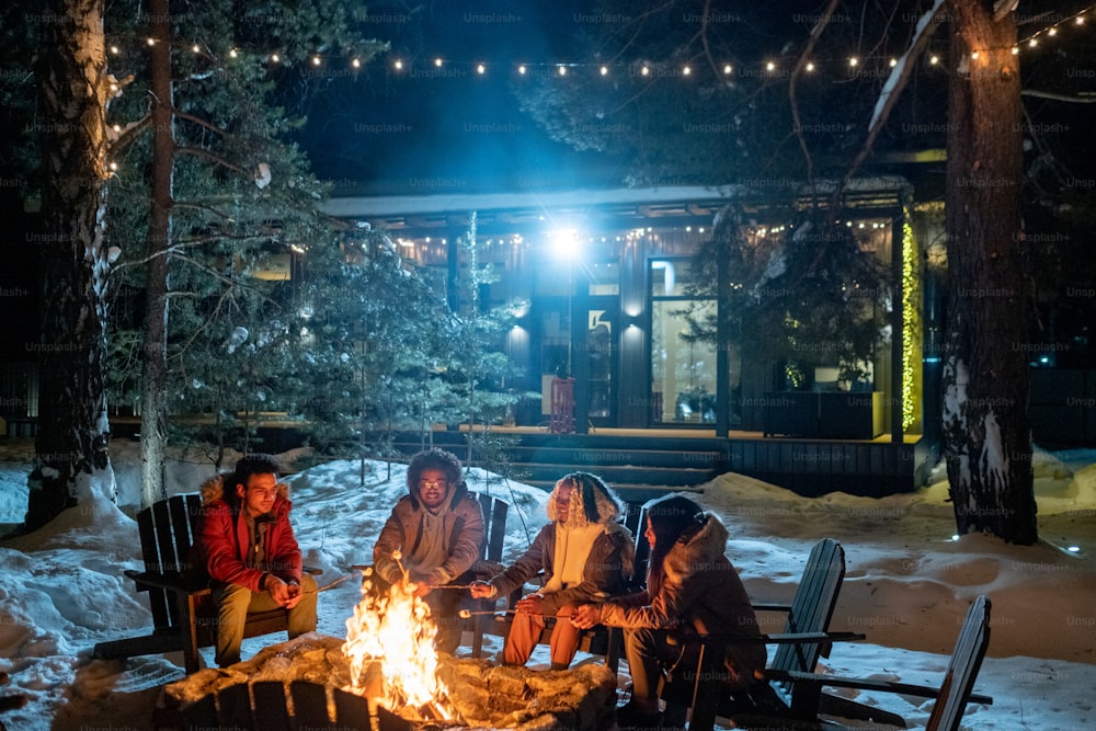 Gruppe von Freunden, die in der Nähe des Feuers im Freien sitzen, Marshmallows kochen und sich bis spät in den Abend während der Winterferien im Landhaus unterhalten