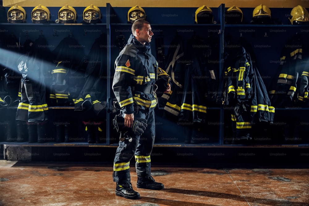 Brave young firefighter standing in fire station in protective uniform and holding helmet under armpit.