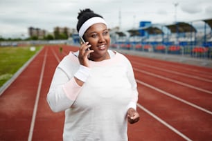 Happy young active woman in sportswear talking by smartphone while training on stadium