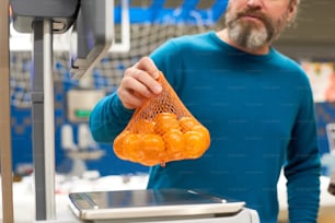Homem maduro com barba grisalha colocando saco com tangerinas frescas em balanças eletrônicas enquanto as pesava no departamento de mercearia