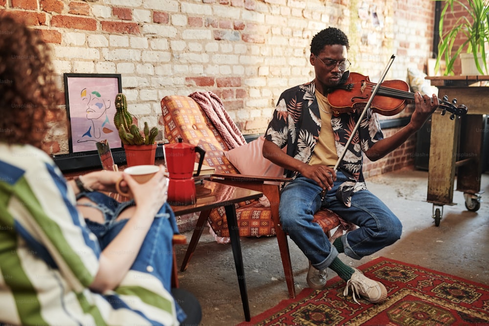 Guy in casualwear of 90s playing violin while sitting in armchair in front of his girlfriend having tea and listening to piece of classic music