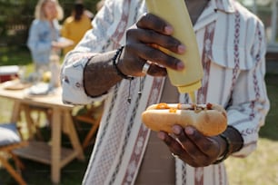 Manos de un joven negro que pone mostaza encima de un perrito caliente con salchicha a la parrilla mientras se prepara un bocadillo durante una fiesta al aire libre