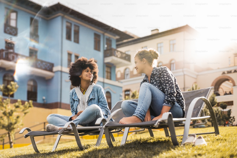 Black girl with curly Afro hair is having a chat with her female smiling Caucasian friend while both sitting on deck chairs on the green fresh lawn between modern houses on a spring sunny day