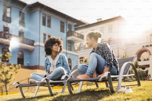 곱슬곱슬한 아프리카 머리를 가진 흑인 소녀는 화창한 날 현대 주택 사이의 녹색 신선한 잔디밭에 있는 갑판 의자에 앉아 웃고 있는 백인 친구와 이야기를 나누고 있다