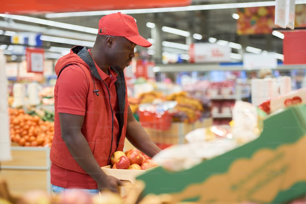 Joven trabajador masculino de un supermercado moderno que lleva una caja de madera con manzanas rojas frescas y la exhibe con frutas