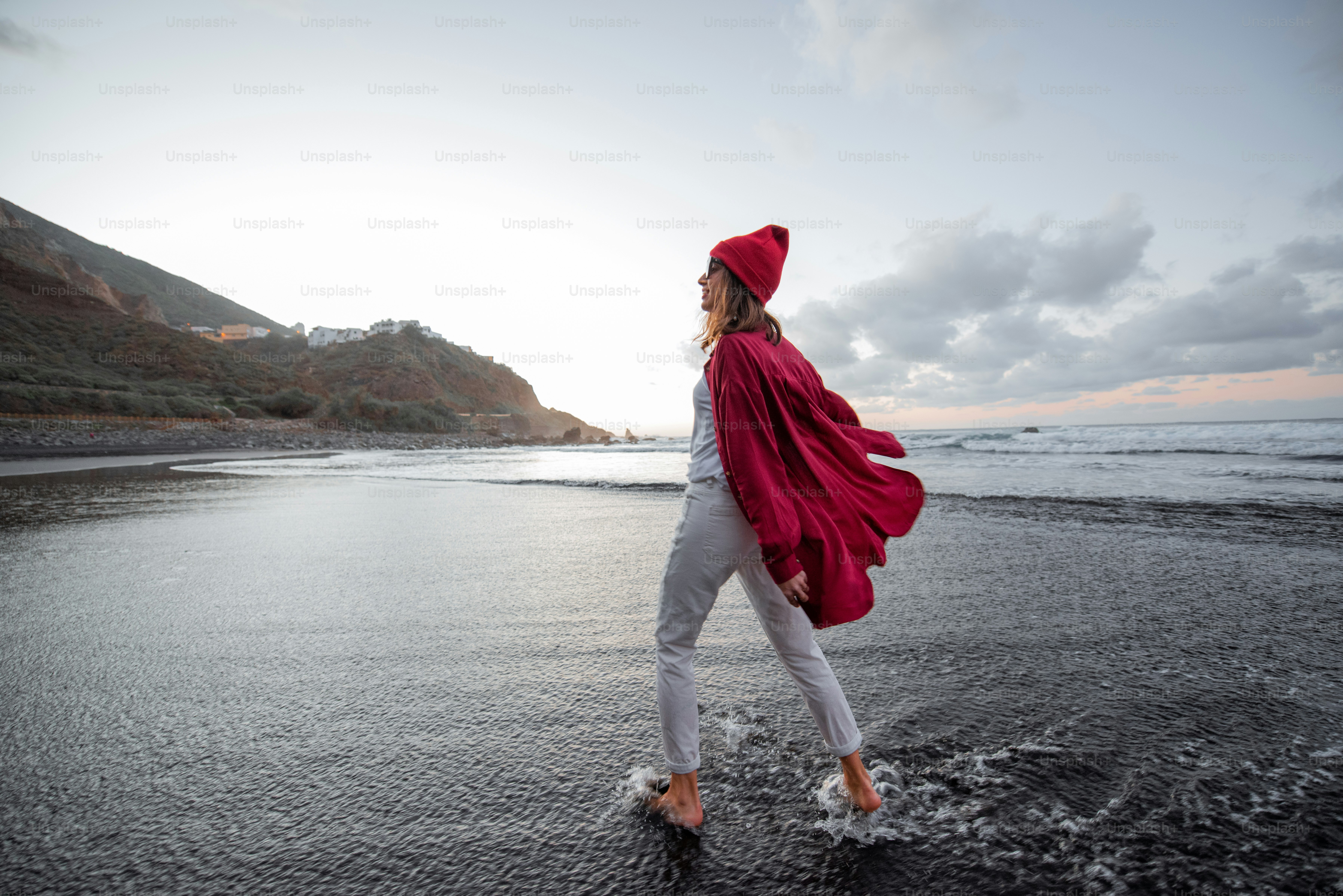 Lifestyle portrait of a carefree woman dressed in red shirt and hat walking on the beach at dusk. Wellness, happiness and life enjoyment concept