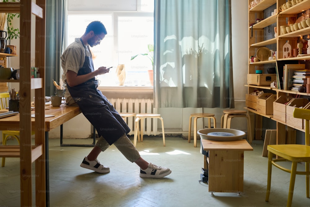 Vista lateral de artesão do sexo masculino jovem em trajes de trabalho rolando no smartphone pelo local de trabalho enquanto olha através de novas ordens de clientes