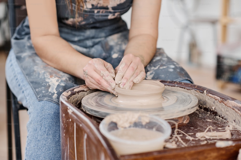 Close-up de jovem artesã criativa habilidosa em cerâmica criando novo item de argila enquanto sentada pela roda giratória na oficina
