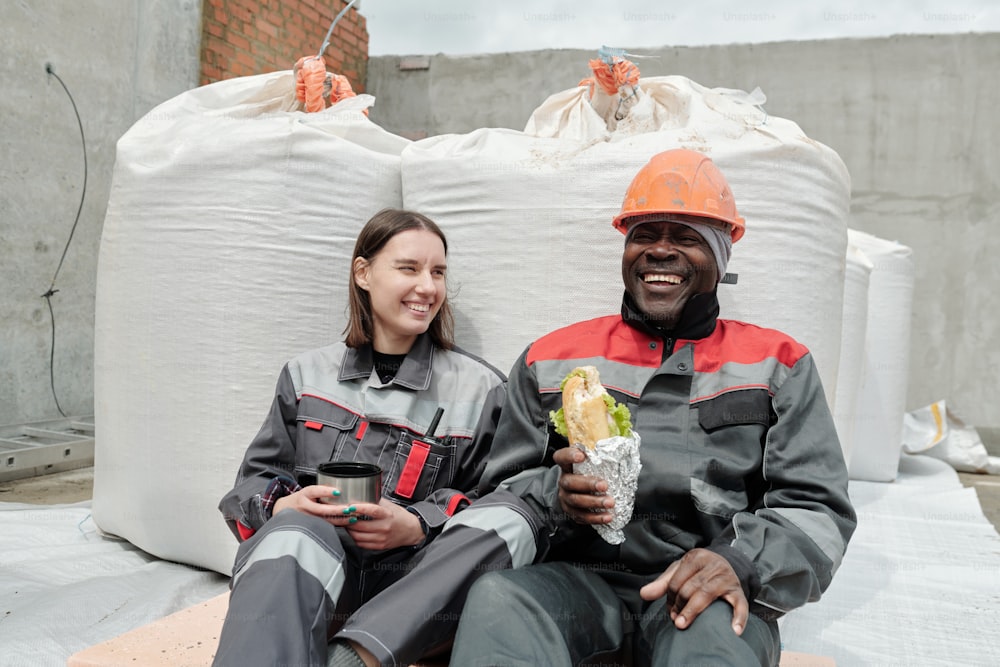 Alegres trabalhadores interculturais do canteiro de obras almoçando contra sacos com concreto ou outros materiais de construção e conversando no intervalo