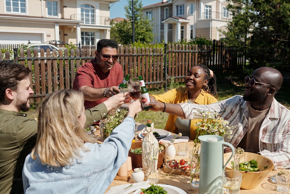 Jovens amigos inter-raciais com copos de vinho caseiro e garrafas de cerveja brindando durante piquenique ou festa ao ar livre sobre mesa servida