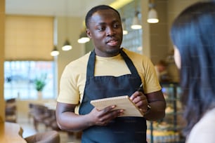 Serveur afro-américain prenant des notes dans un bloc-notes tout en parlant à une femme dans un café, il prend sa commande