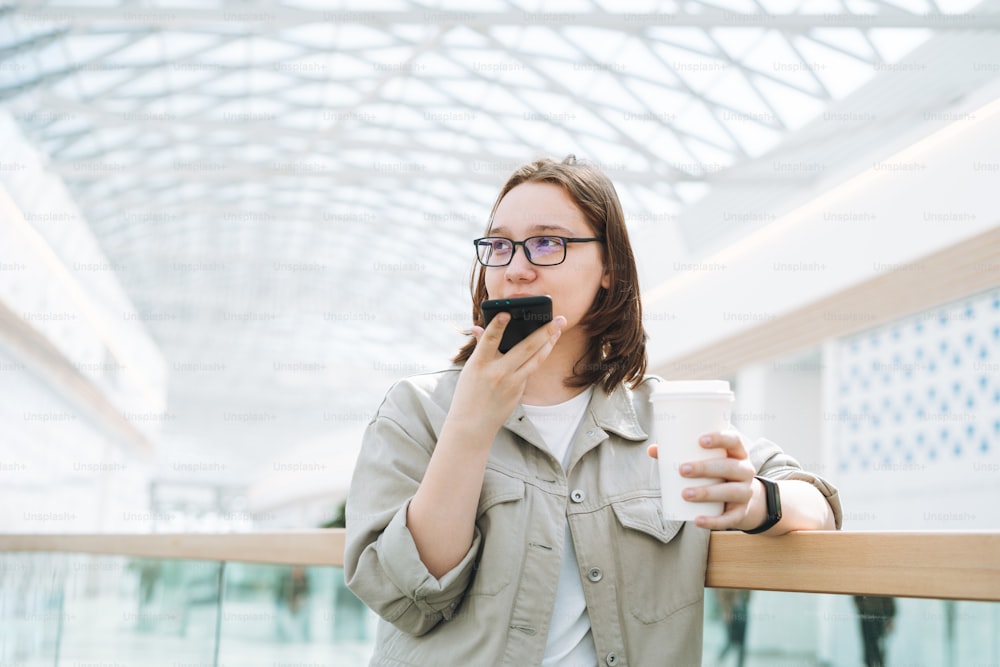 Giovane studentessa adolescente bruna in occhiali usando il telefono cellulare ha inviato un messaggio vocale con tazza di caffè di carta al centro commerciale del luogo pubblico