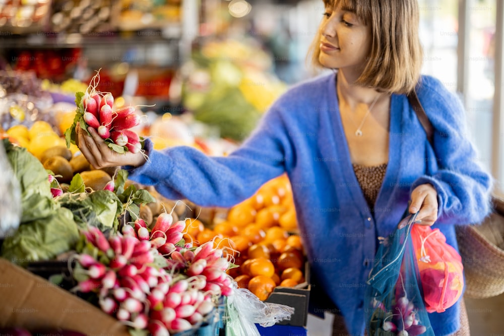 Une femme prend un bouquet de radis sur le comptoir, achète des légumes et des fruits frais au marché local. Faire ses courses avec un sac en filet réutilisable. Durabilité et concept d’alimentation biologique