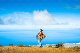 Overjoyed woman tourist opening and outstretching arms in front of a blue sea and sky scenic beautiful background. Happy travel lifestyle for female people wearing colorful clothes