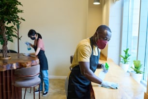 カフェで感染コロナウイルスを保護するための消毒スプレーを備えたマスククリーニングテーブルのアフリカのウェイターと背景の同僚