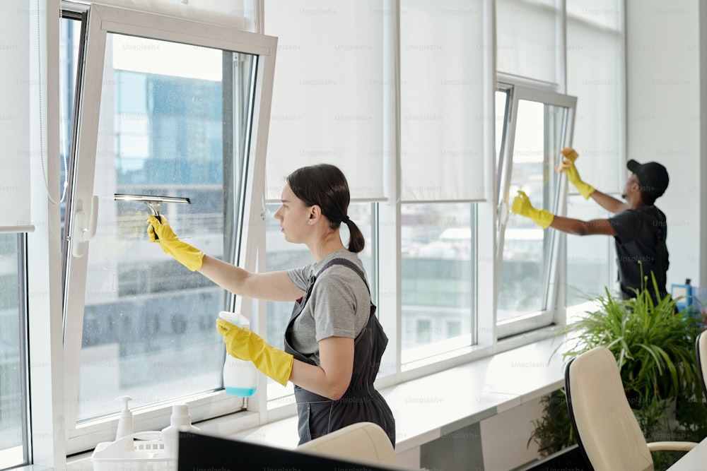 Young female cleaner in workwear washing large windows of openspace office against her African American male colleague