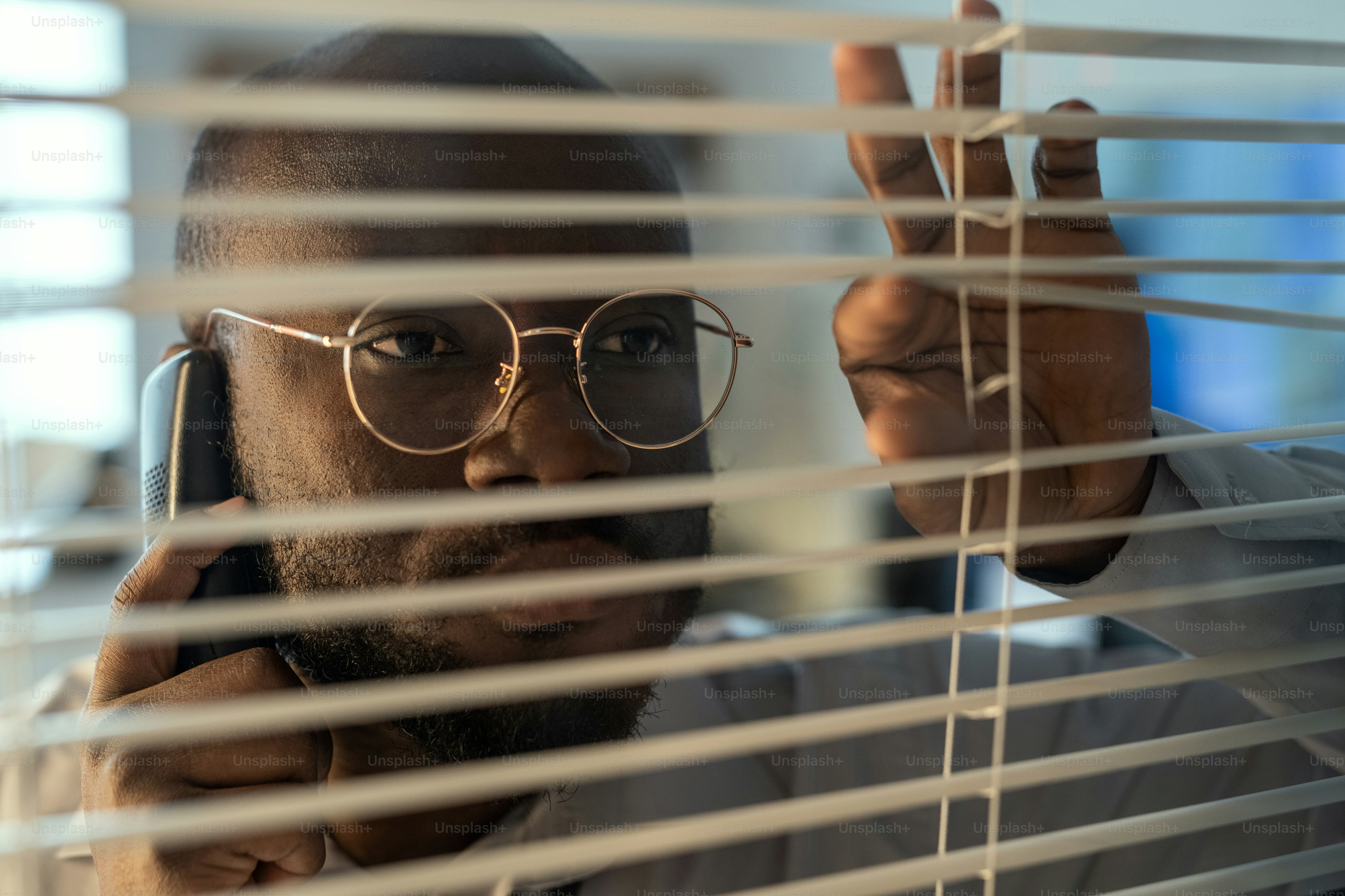 Volto dell'ufficiale afroamericano in occhiali che parla al telefono cellulare mentre guarda attraverso le tende veneziane nel dipartimento di sicurezza