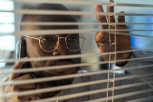 Face of African American officer in eyeglasses speaking on mobile phone while looking through venetian blinds in security department