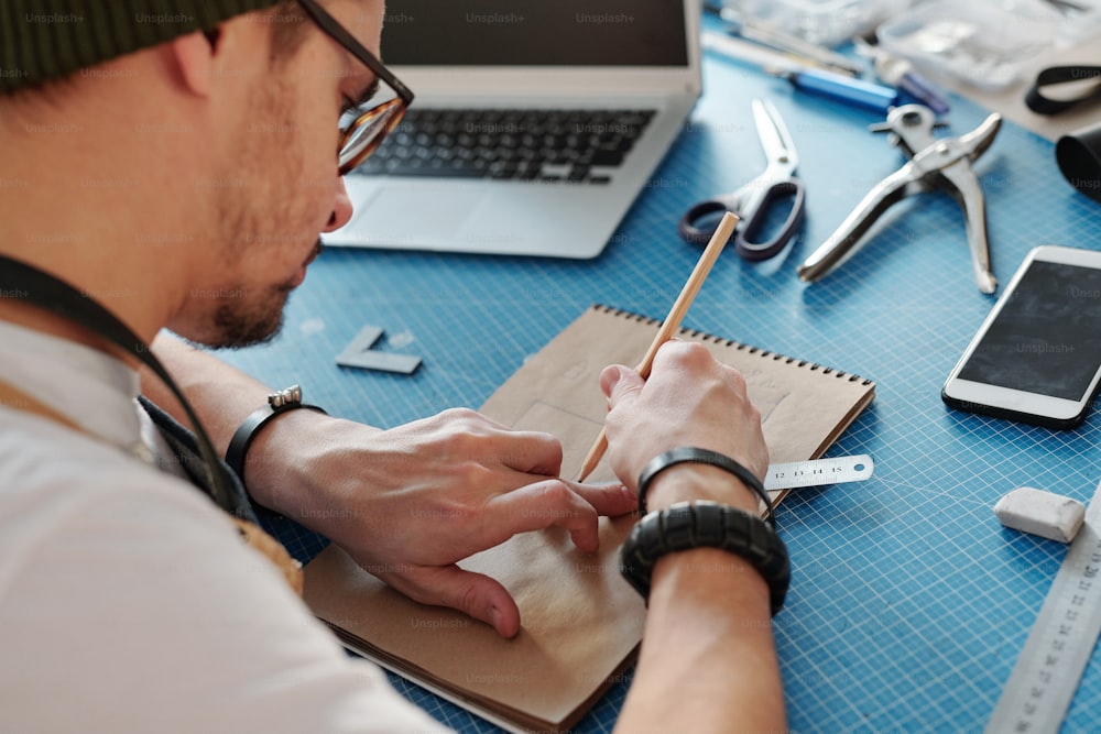 Über die Schulter Blick auf einen beschäftigten jungen Mann, der das Design eines Lederprodukts am Tisch zeichnet