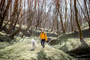 Young woman in hiking clothes and backpack spend time together with big white dog in green spring forest. Enjoys and explore of tranquil nature.