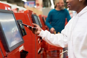 Jovem negra com smartphone fazendo pagamento sem contato por terminal de autoatendimento enquanto paga por produtos alimentícios no supermercado
