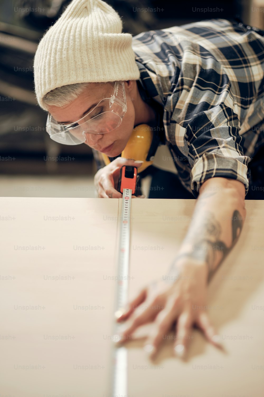Ritratto di donna tatuata sui 40 anni che lavora in falegnameria con occhiali protettivi, misurando una tavola di legno. Occupazione maschile, lavoro maschile, concetto di uguaglianza di genere