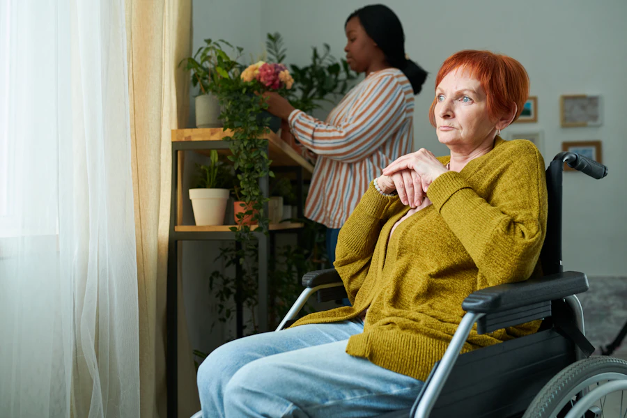 An older woman seated by a window while a carer arranges flowers behind her