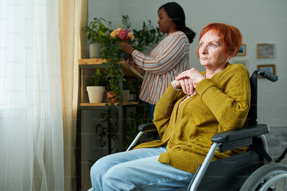Elderly woman sitting on wheelchair and looking at window with pensive expression with caregiver caring of plants in background