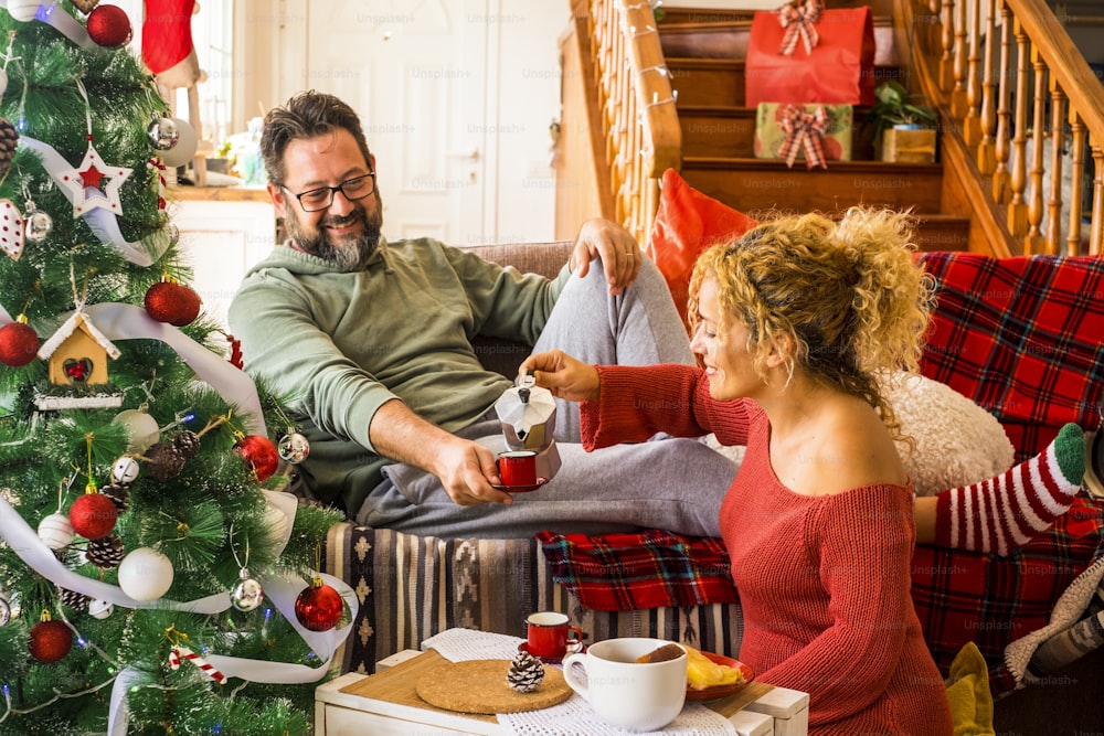 Christmas time at home with happy adult caucasian couple having breakfast on the couch near the christmas tree - concept of family and love life together in traditional leisure activity indoor