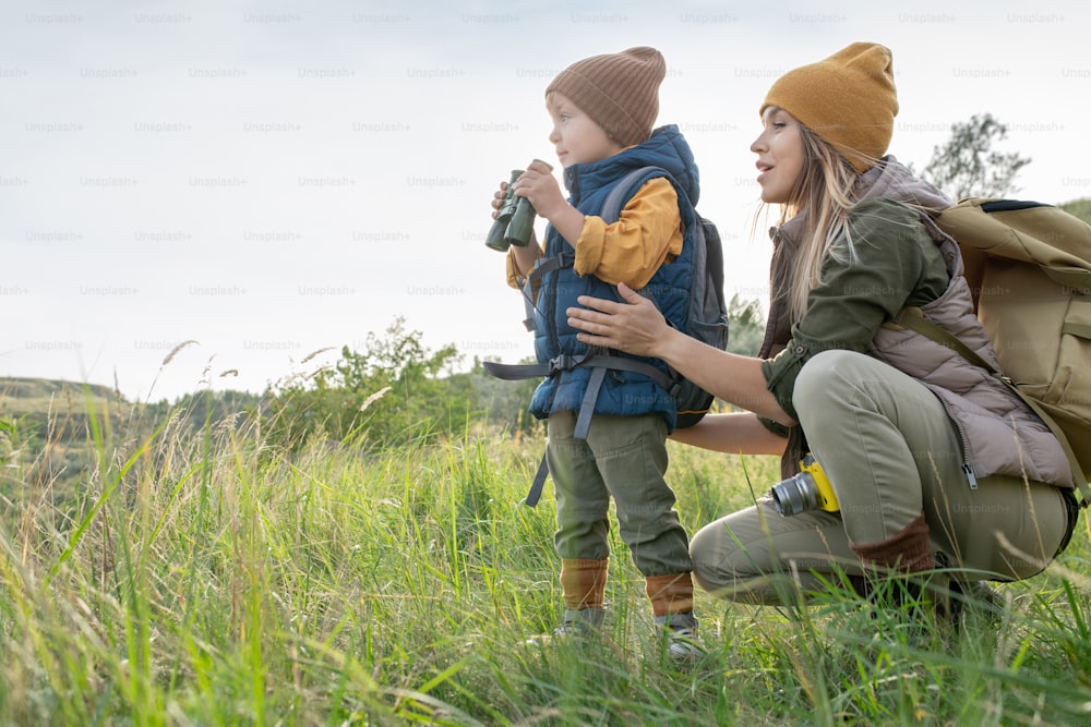 Young woman with backpack squatting by her little son with binoculars during weekend trip in natural environment among green grass