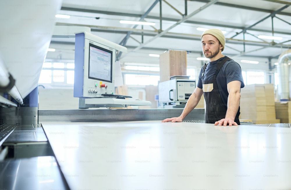 Young worker standing at cutting machine to work with big wooden board at furniture factory