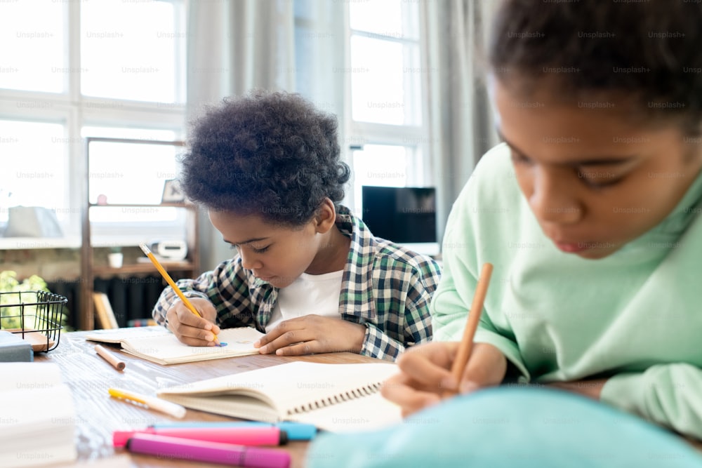Lindo niño de primaria con ropa informal y su hermana tomando notas en cuadernos mientras hace la tarea en la mesa en el entorno del hogar