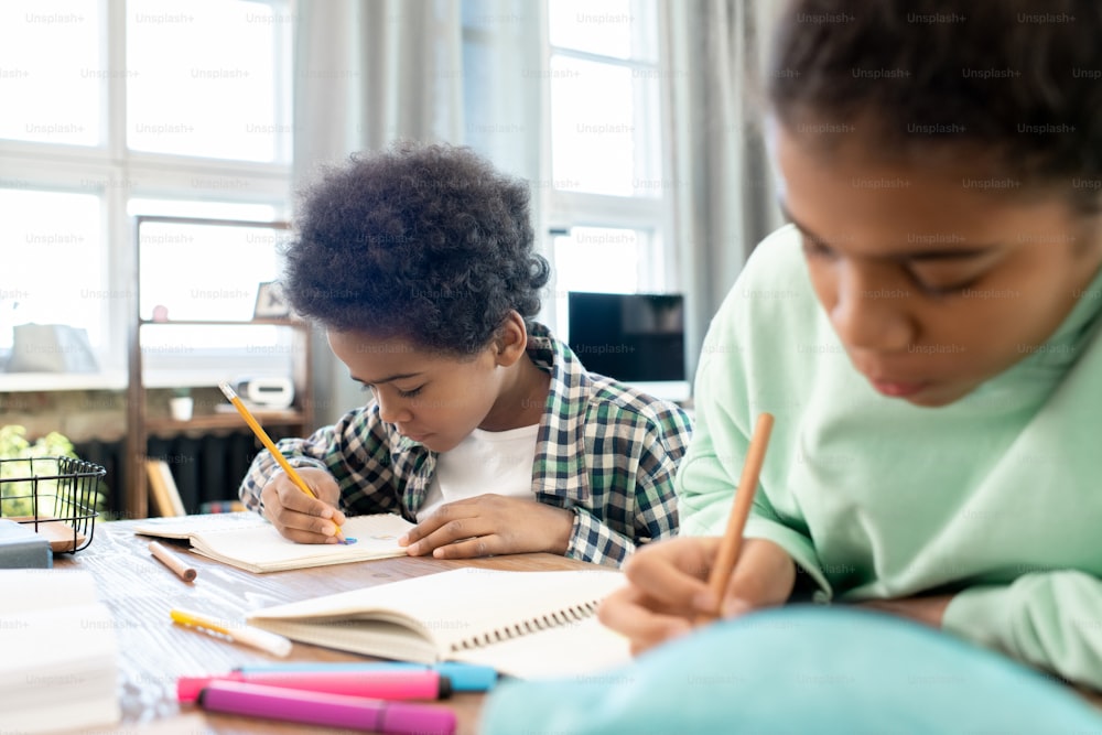 캐주얼 차림의 귀여운 초등학생과 그의 여동생은 가정 환경에서 테이블 옆에서 숙제를 하면서 카피북에 메모를 하고 있다