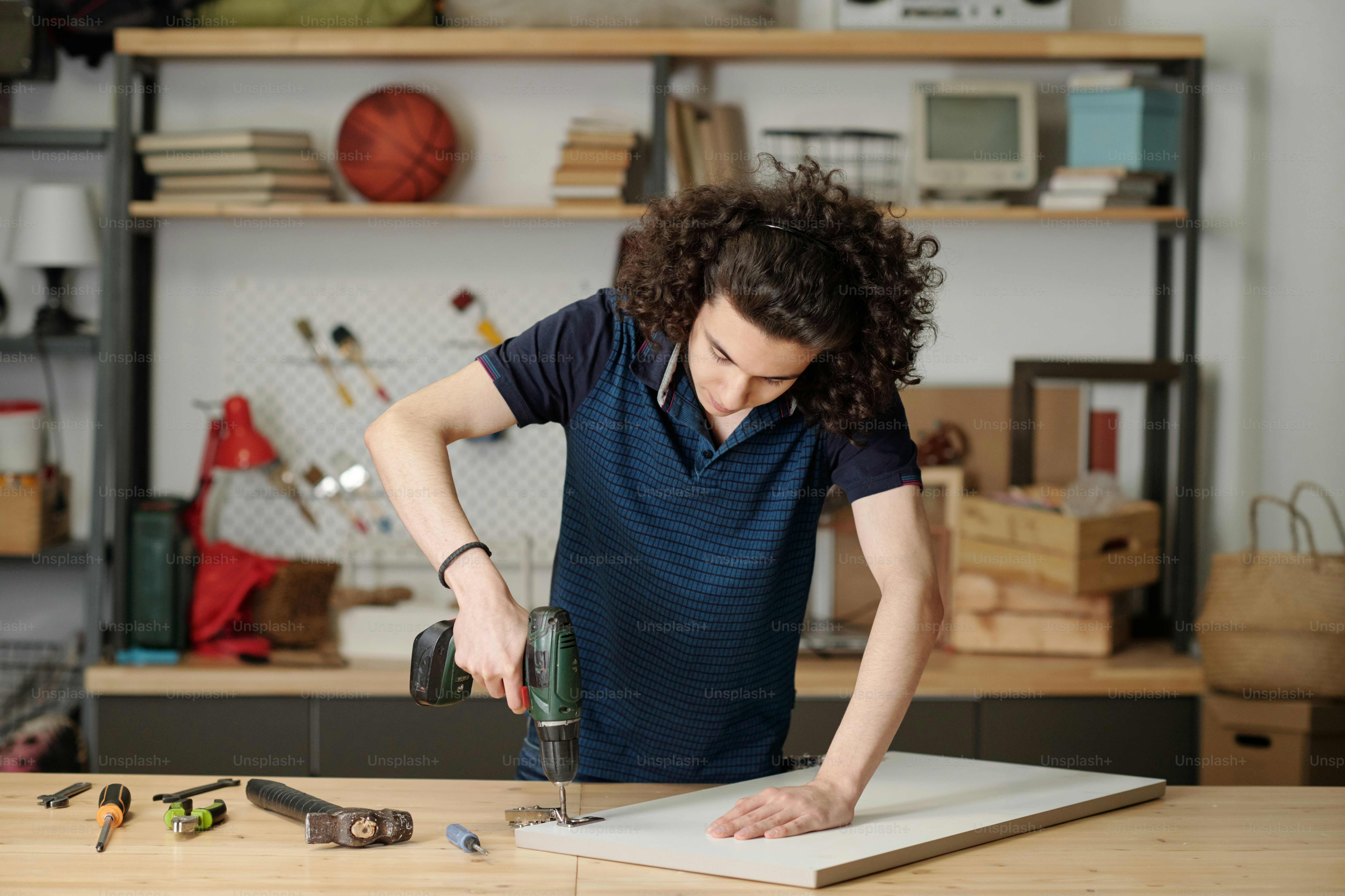 Teenage boy in blue casual shirt using electric drill while repairing plywood door of kitchen cabinet in garage of his house