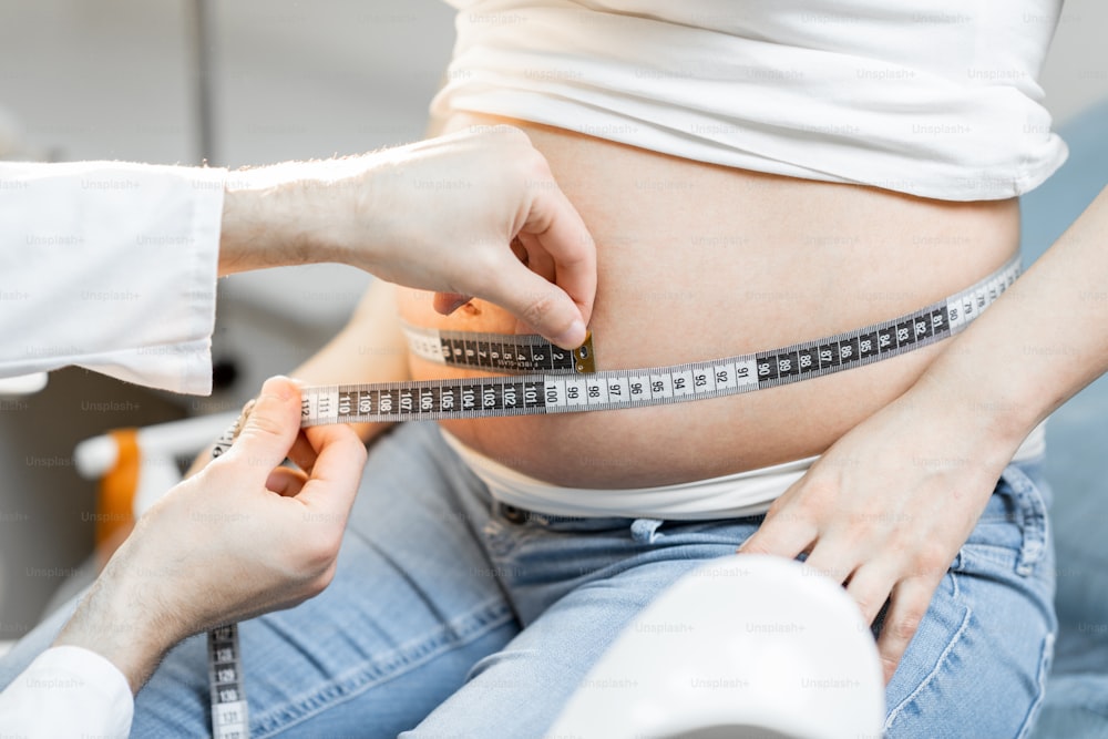 Doctor midiendo el vientre de la mujer embarazada con una cinta durante un examen médico, vista recortada sin rostro centrado en el abdomen