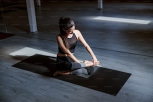 Linda morena em traje ativo colocando a perna em posição de lótus. Interior do estúdio de ioga.