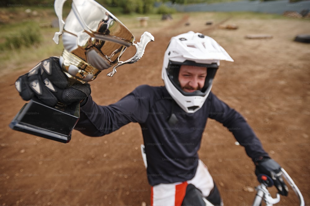 Vue ci-dessus d’un motocycliste heureux en gants levant la main avec la coupe gagnante tout en montrant le prix pour la compétition sur route