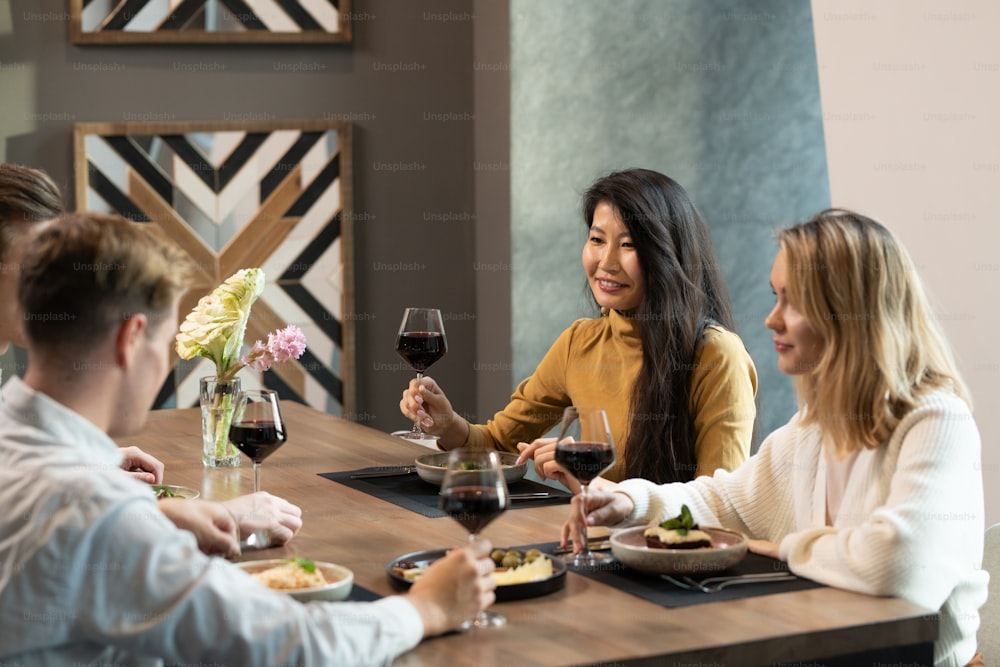 Joven elegante mujer asiática con una copa de vino tinto haciendo brindis por la cena mientras está sentada junto a la mesa servida en el restaurante entre sus amigos